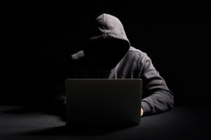 A suspicious figure in a hoodie at a table with a laptop.